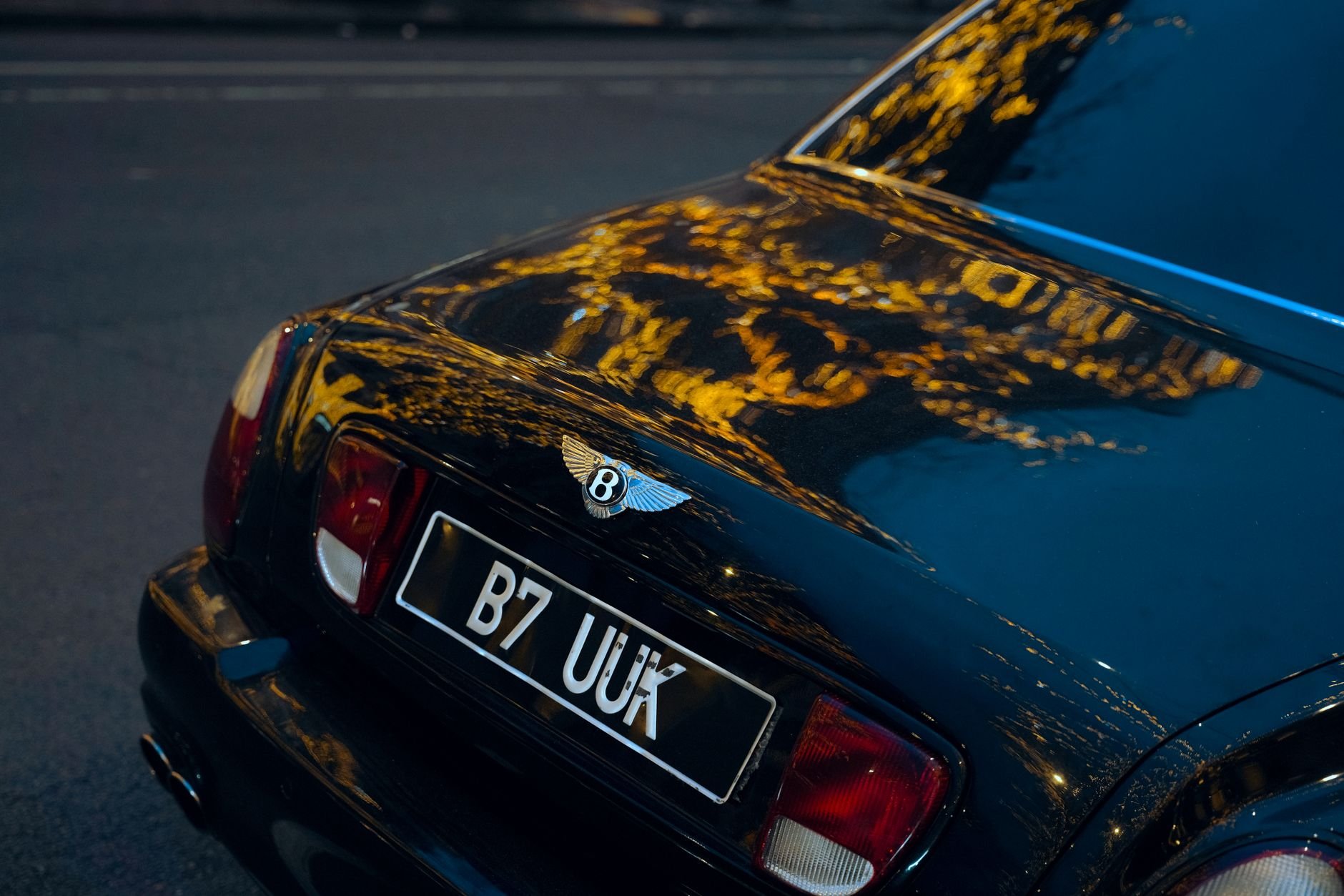 Close-up of a luxury car's rear with city lights reflecting on dark paintwork at night.